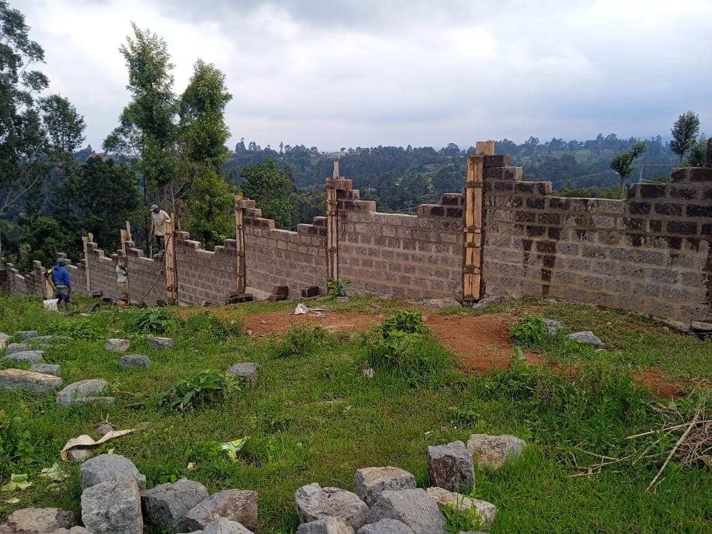 Block wall with wooden formwork supports for pillars, worker visible on left, stones in foreground with scenic valley view