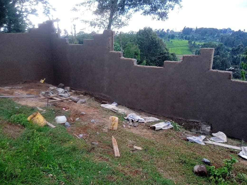 Corner section of plastered wall with battlement top, construction materials and yellow containers on ground, green hills in background