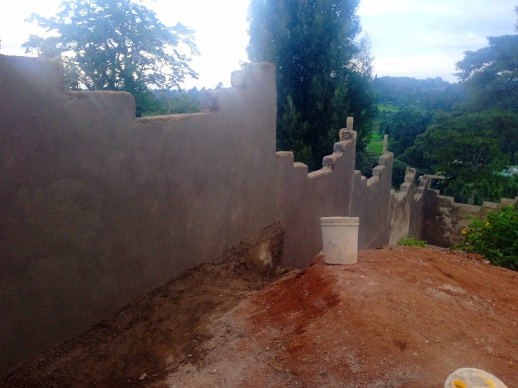 Plastered perimeter wall with pillars under construction, bucket and red soil visible with scenic green landscape