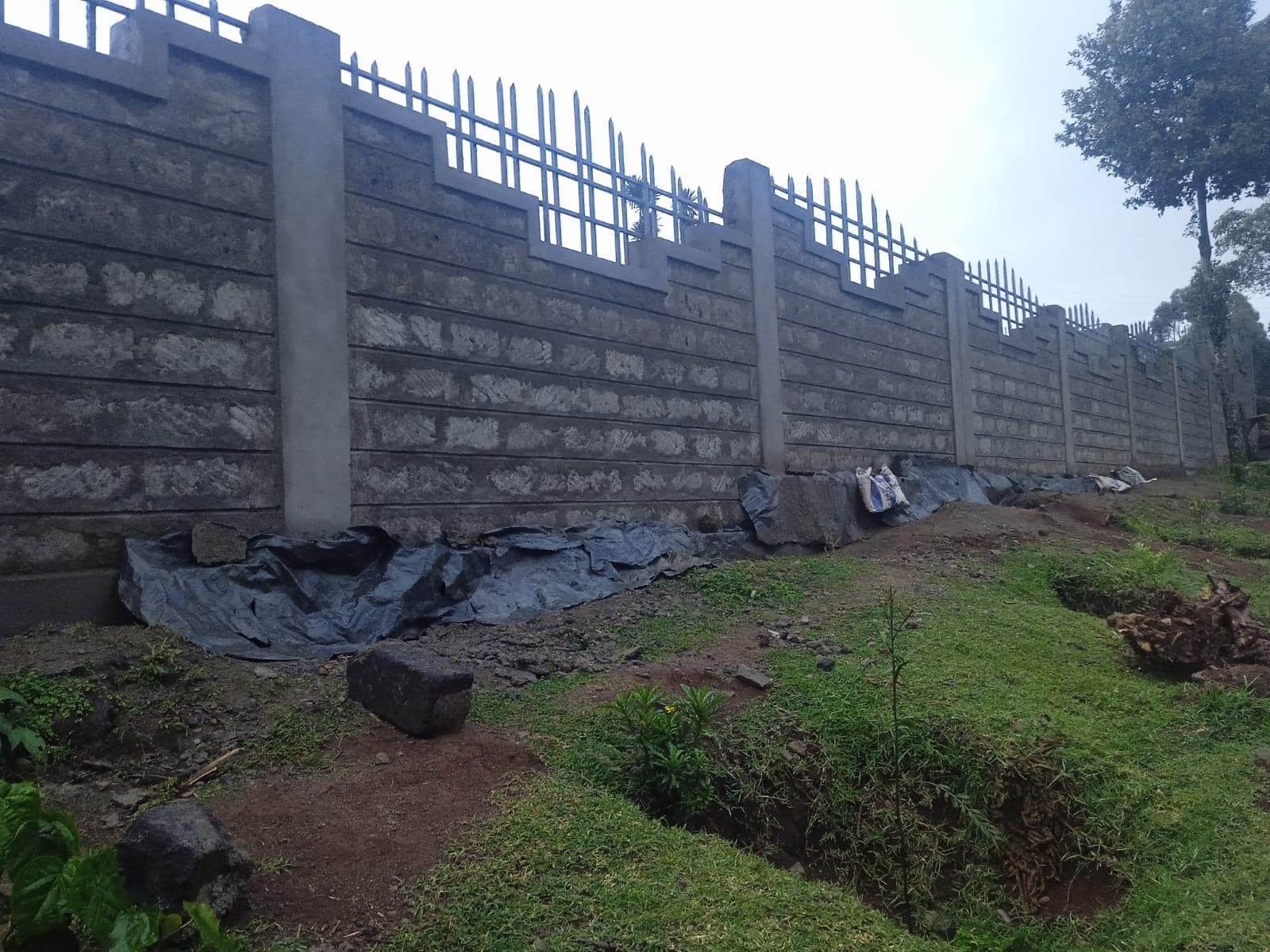 Tall wall section with grooved block texture, metal railings, black tarp at base for foundation protection, grassy surroundings
