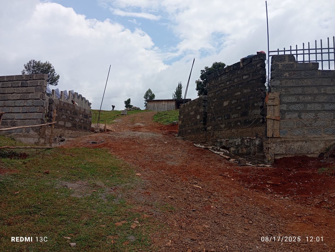 Main entrance gate area with stone block pillars on either side, metal railings visible, red soil path leading uphill