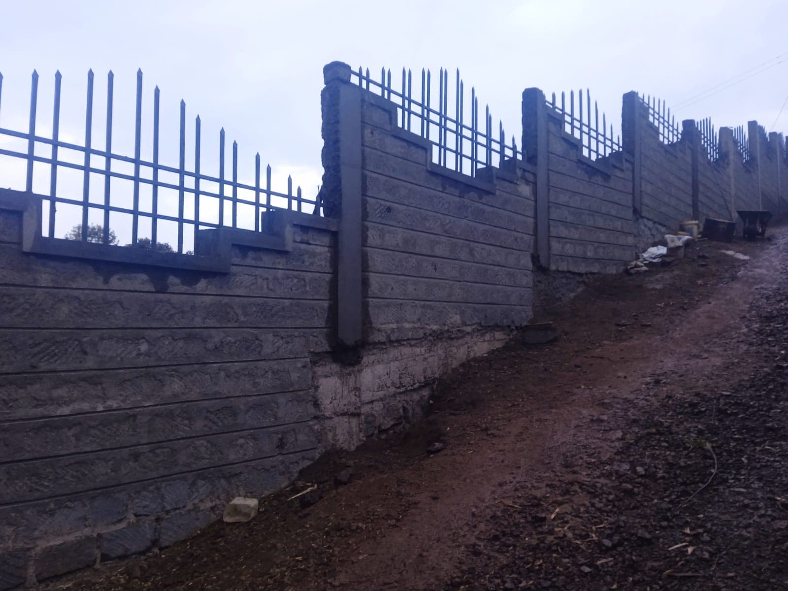 Exterior view of completed grooved-texture perimeter wall with metal security railings, decorative stepped pattern, dirt path alongside