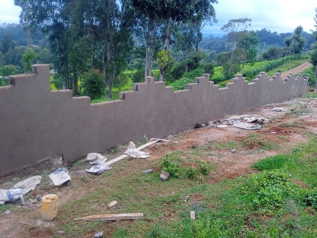 Long plastered perimeter wall with decorative crenellated battlement top, construction debris at base, lush green valley backdrop