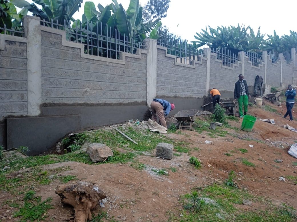 Multiple workers doing finishing work at base of wall with wheelbarrow and green bucket, banana trees behind decorated wall