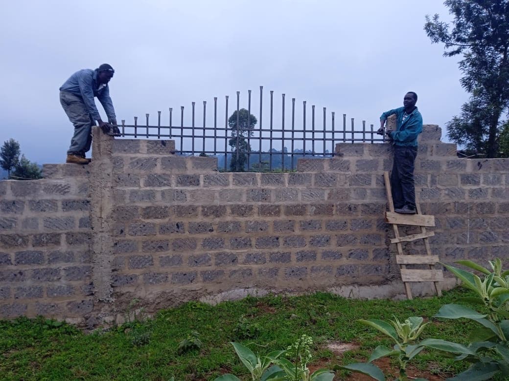 Two workers on top of block wall installing metal railings, wooden ladder against wall, grassy site