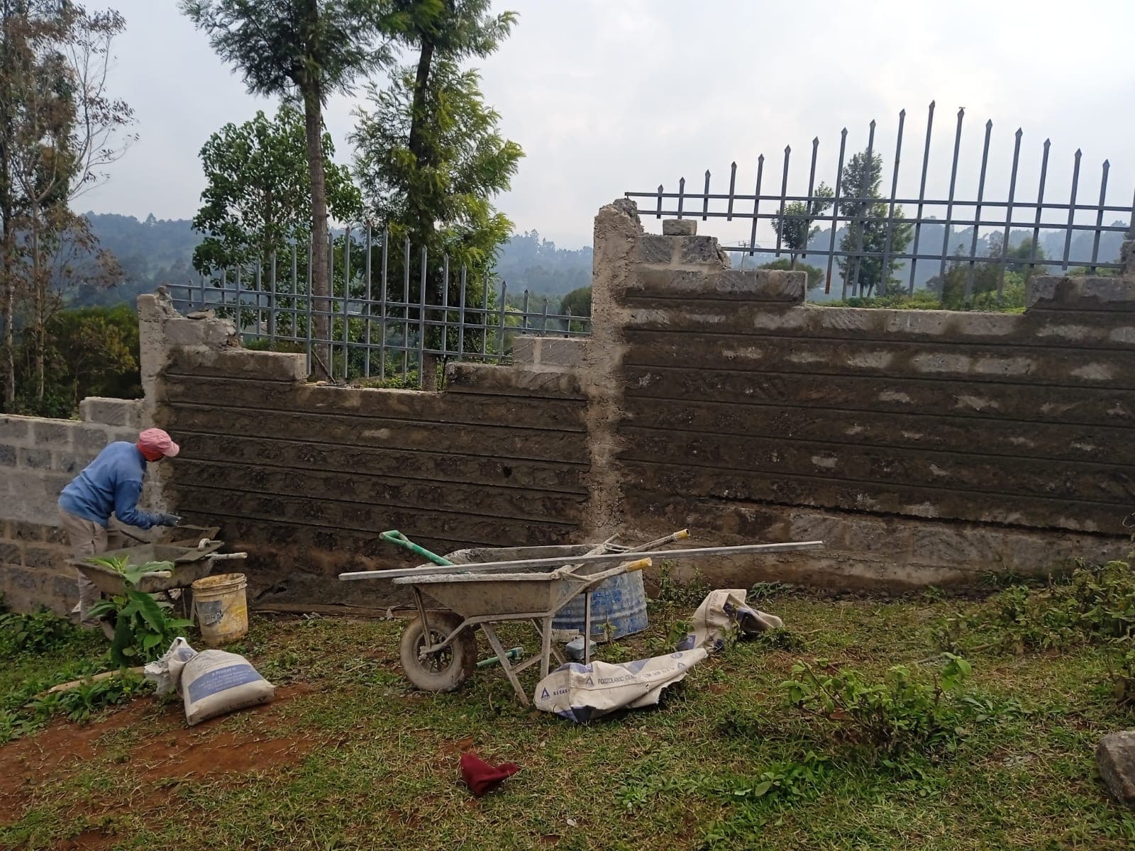 Worker in red cap plastering grooved texture wall with wheelbarrow and cement bags, metal railings on top