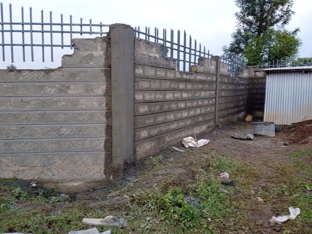 Corner section of grooved-texture block wall with metal railings, plastered pillar, corrugated metal storage shed in background